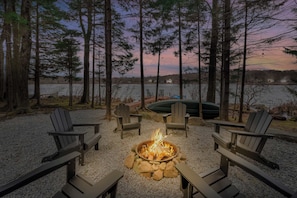 A fire pit surrounded by Adirondack chairs, a gravel area, and a lake with boats and houses in the background. - Lakefront Hot Tub Kayaks High End Peaceful (Tobyhanna)