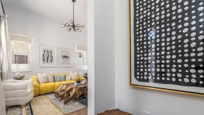 Una sala moderna con un sofá amarillo, un sillón blanco y una mesa de centro de madera. Hay cuadros enmarcados en la pared y una obra de arte grande con lunares.