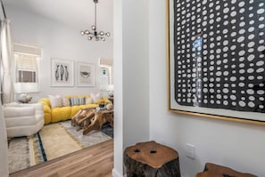 Ein modernes Wohnzimmer mit einem gelben Sofa, einem weißen Sessel und einem Holztisch. An den Wänden befinden sich gerahmte Bilder und ein großes Kunstwerk mit Punkten.