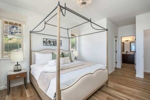 Ein Bett mit Baldachin in einem gut beleuchteten Zimmer mit Holzboden, einem Nachttisch mit Lampe und einem gerahmten Bild an der Wand.