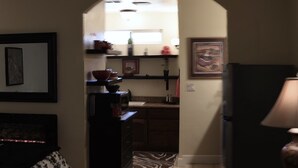 Ein Wohnzimmer mit einer Couch, einem Couchtisch, einer Lampe und einer Küchenzeile mit Kühlschrank, Mikrowelle und Regalen.
