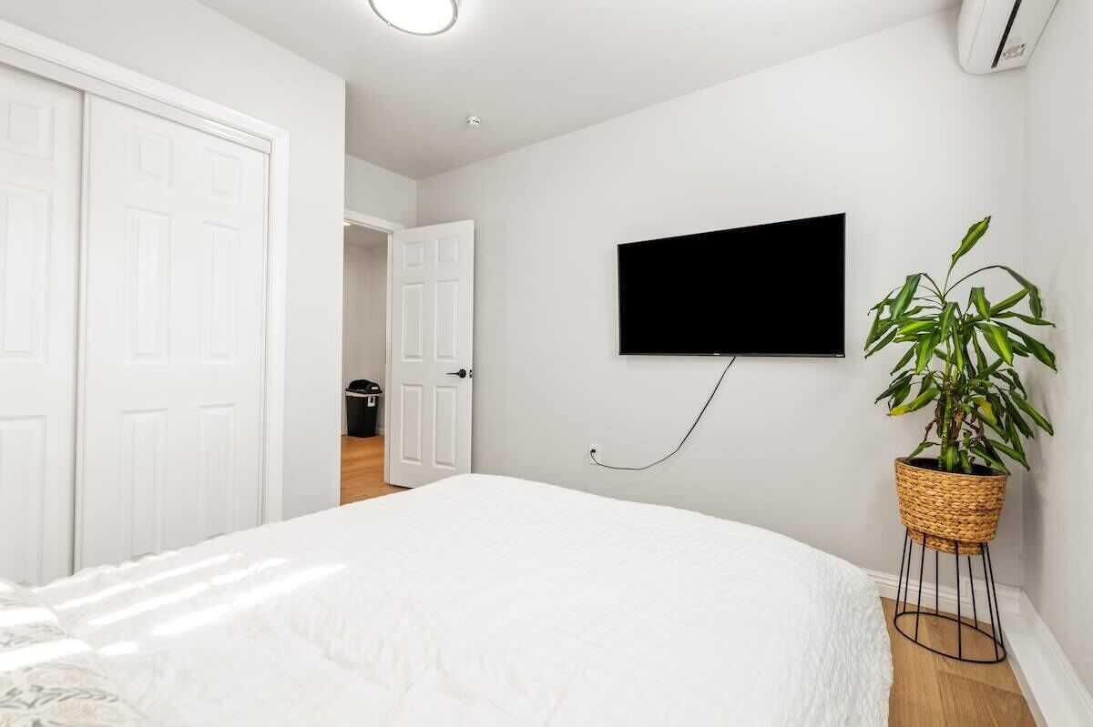 A bedroom with a bed, a TV mounted on the wall, a potted plant, and a door leading to another room.