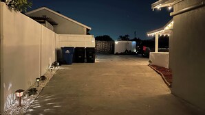 En asfalterad uppfart leder fram till en byggnad med vit fasad och en garageport. Längs uppfarten står tre soptunnor och en ljusslinga är upphängd.