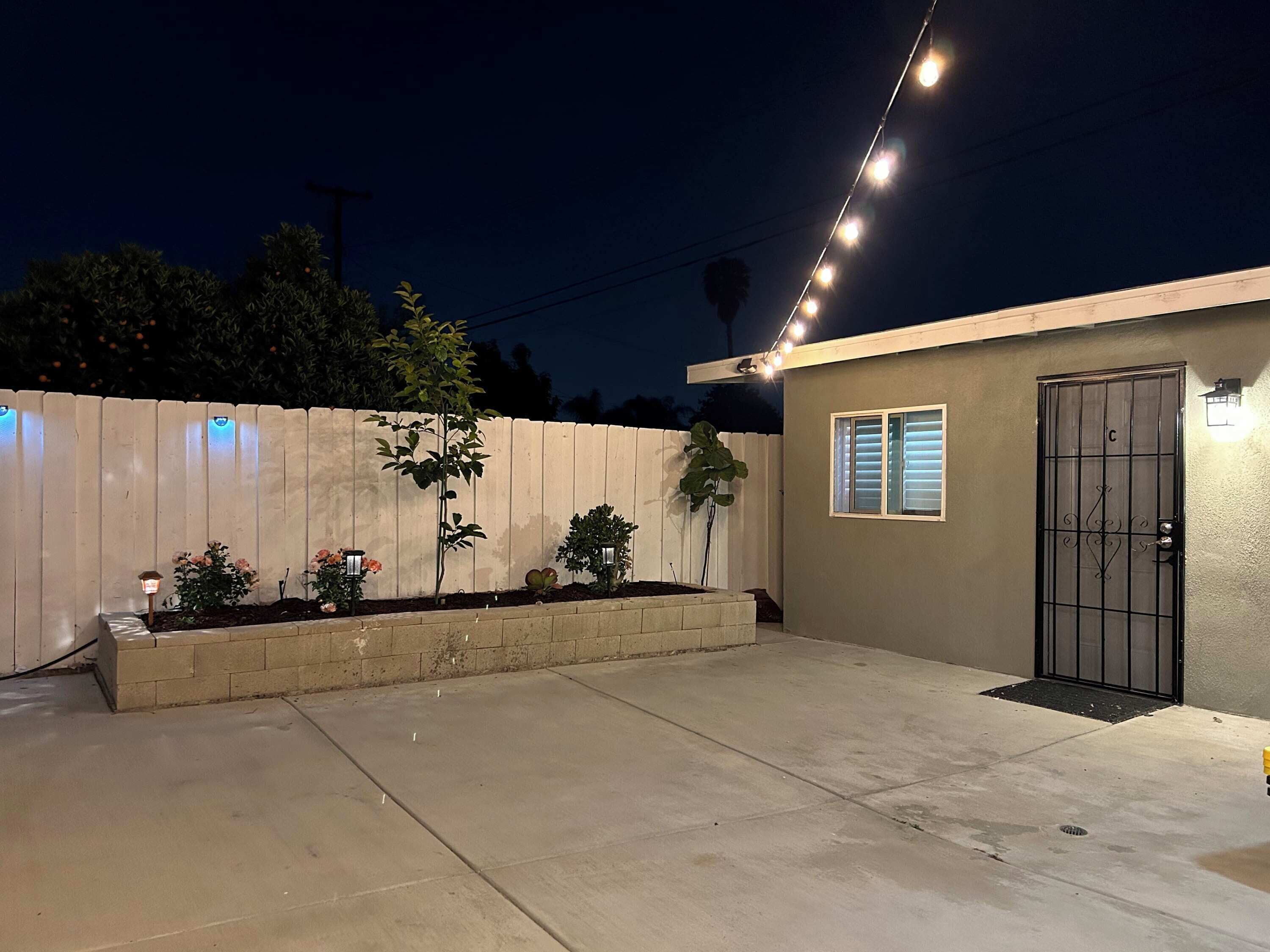 A residential area with a concrete patio, a white fence, and a small building with a metal gate.