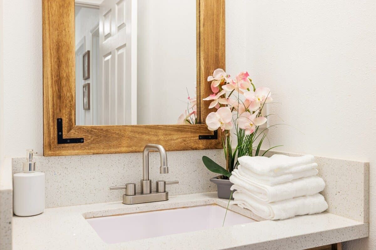 Une salle de bain avec un miroir à cadre en bois, un robinet moderne, un lavabo, une orchidée en pot et une pile de serviettes blanches.