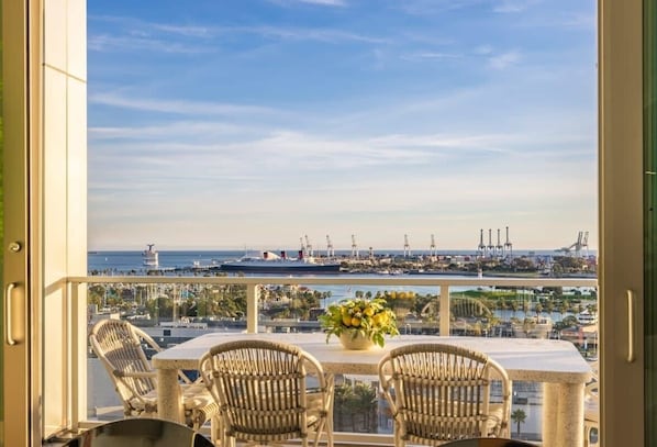 Un balcon agrémenté de fauteuils et d’une table en osier, donnant sur une marina où sont amarrés des bateaux et où se dressent des structures industrielles.