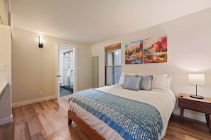 A bedroom with a bed, a nightstand, a painting, and a door leading to a bathroom.