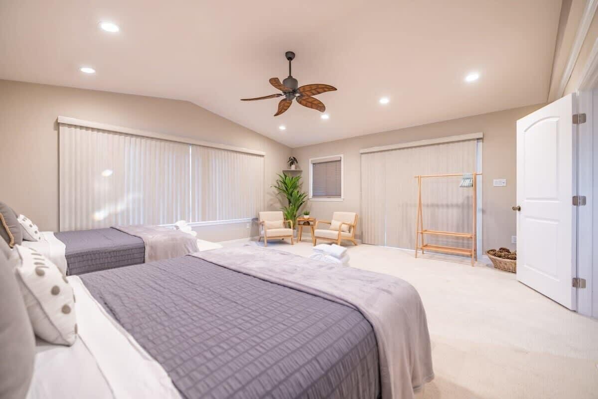 A bedroom with two beds, a ceiling fan, a window with blinds, a small table with chairs, and a door.