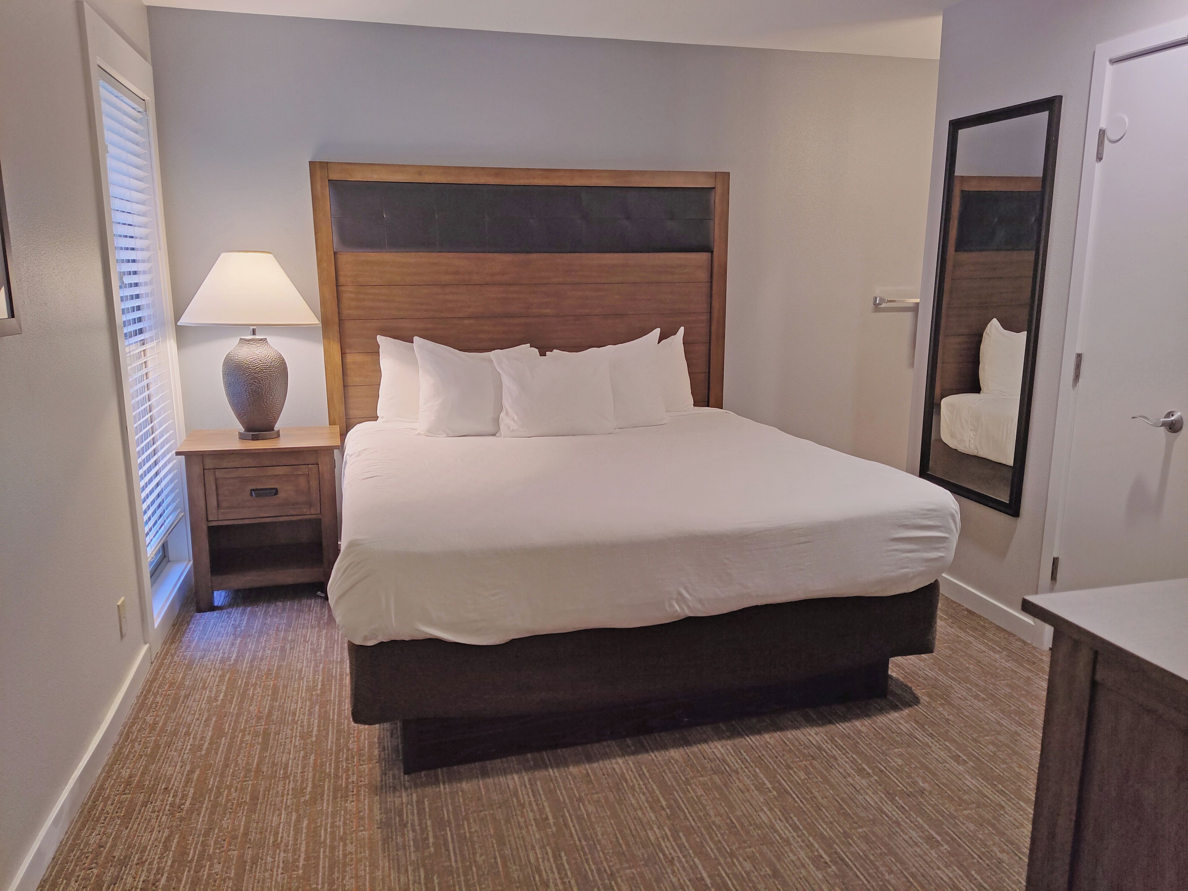 A neatly made bed with white linens, a wooden headboard, a bedside table with a lamp, and a mirror on the wall.