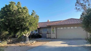 A residential house with a garage, a driveway, and a large tree in the front yard. - @ Marbella Lane - Majestic Desert Mountain Views (Desert Hot Springs)