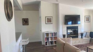 A living room with a sofa, bookshelf, flat-screen TV, fireplace, and a dining area. - @ Marbella Lane - Majestic Desert Mountain Views (Desert Hot Springs)