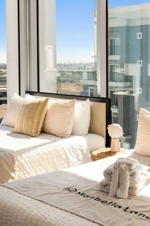 A hotel room with a large window, a bed with multiple pillows, a bedside table with a lamp, and a towel on the bed. - @ Marbella Lane - Panoramic Delight Dream Escape! (Long Beach)
