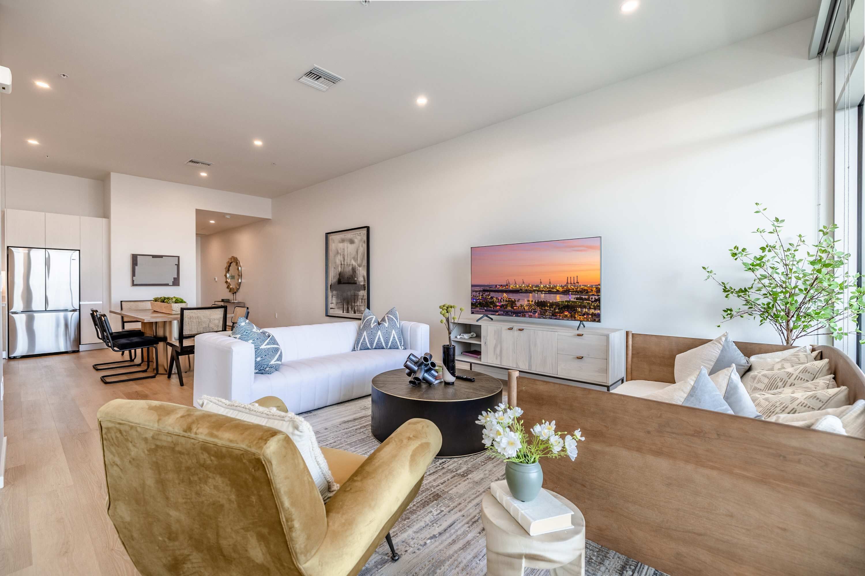 Una sala moderna con sofá, sillón y mesa de centro, un área de comedor con sillas y mesa, y un ventanal con vista a la ciudad.
