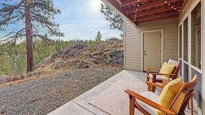 A covered patio with two wooden chairs and a rug, overlooking a rocky terrain and trees. - Seventh Mountain 1417 (Bend)