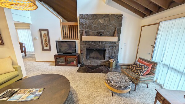 A cozy living room with a stone fireplace, a television on a wooden stand, and a comfortable seating area with a patterned ottoman and cushions.