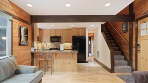 A modern kitchen with wooden cabinets, a central island, and a microwave. There is a staircase leading to a loft area. - Pole House 10 (Bend)
