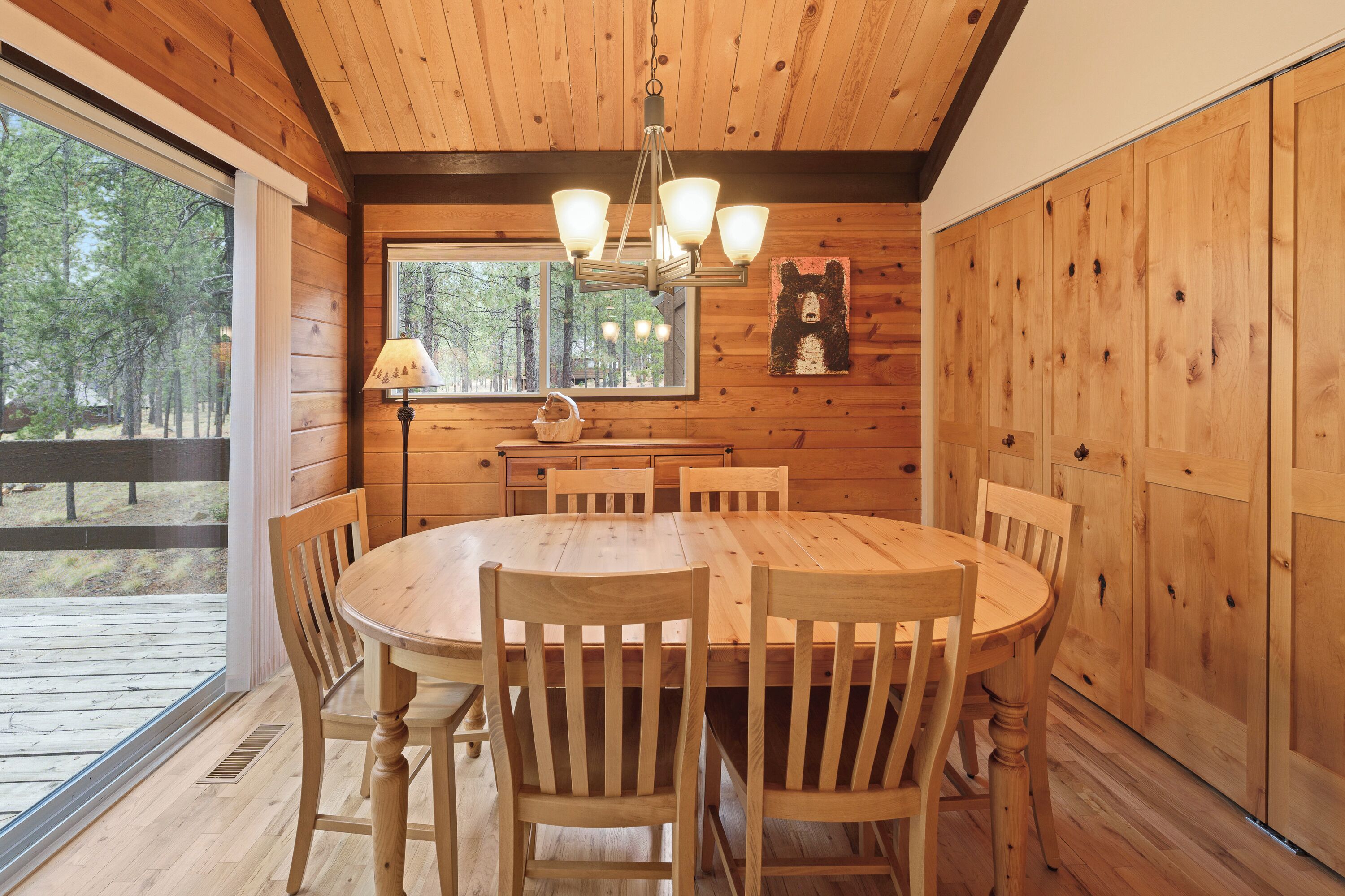 Une salle à manger en bois avec une grande table ronde et des chaises, un lustre et une fenêtre donnant sur les arbres.