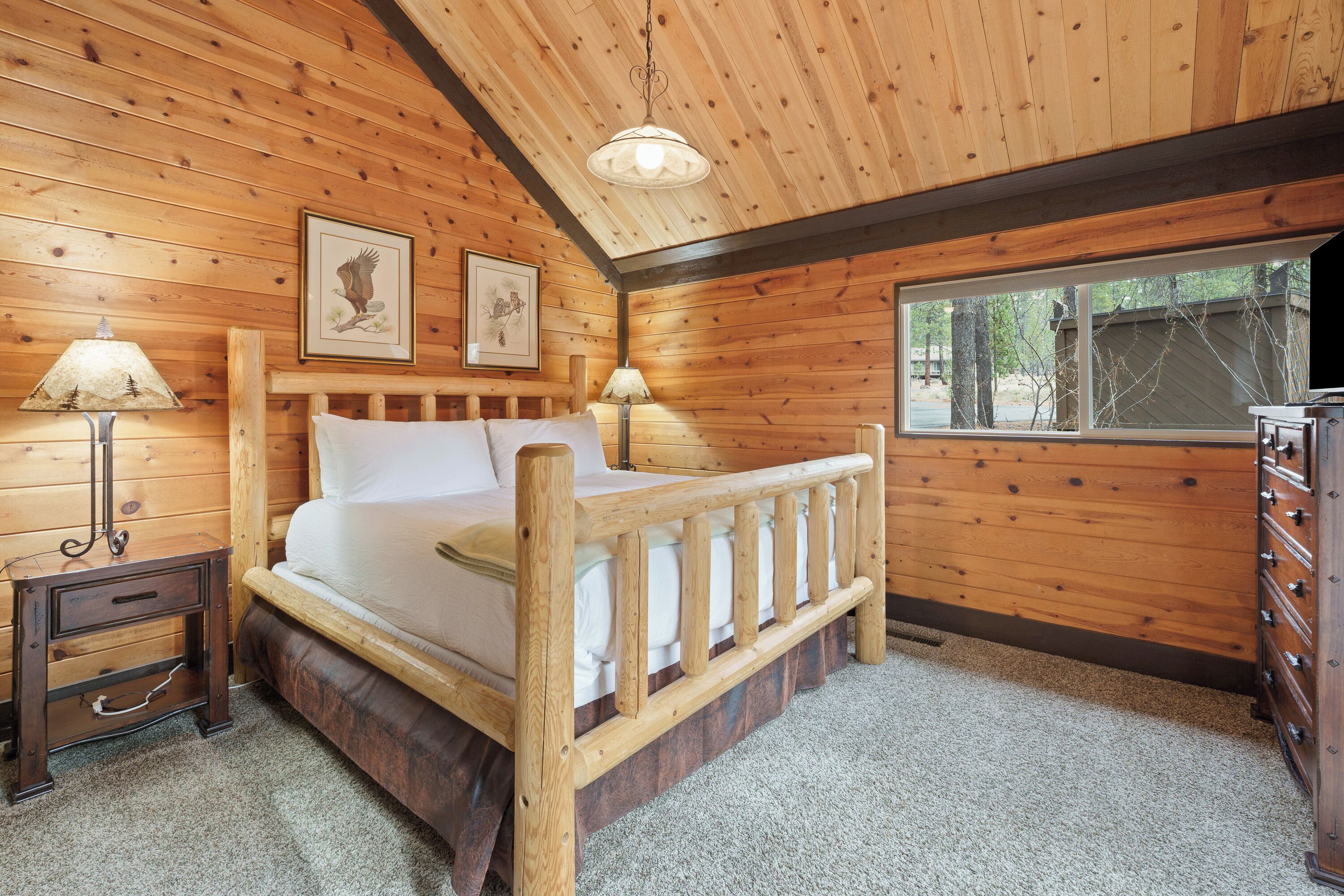 A wooden cabin bedroom with a bed, two lamps, a dresser, and framed pictures on the wall.