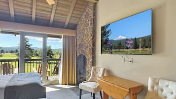 A room with a bed, a wooden table, a white sofa, a chair, a stone wall, a ceiling fan, and a large window with a mountain view.