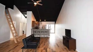 Un interior moderno con suelo de madera, un sofá de cuero negro, un mueble para televisión y una zona de cocina con taburetes de bar.
