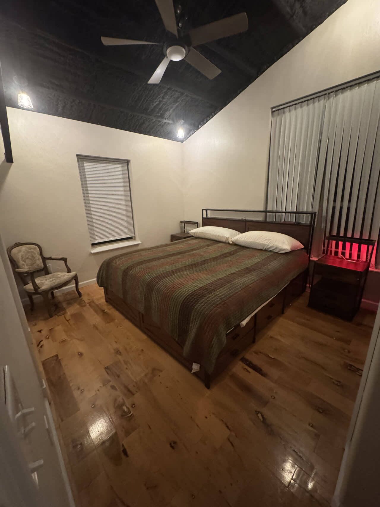 Een slaapkamer met een bed, een stoel, een plafondventilator, een raam met jaloezieën en een rood licht.