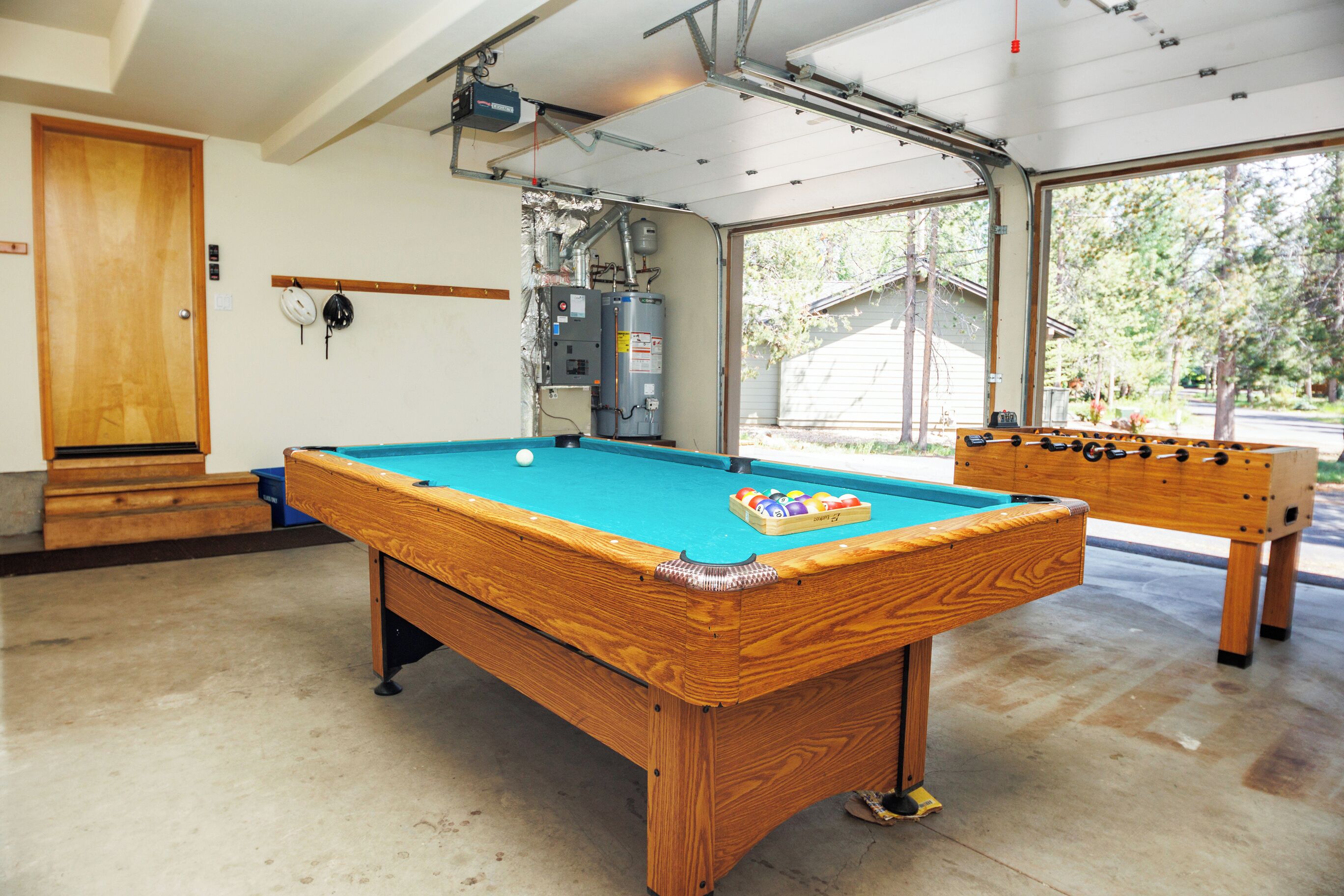 Une table de billard recouverte d’un drap bleu, une table de baby-foot et une grande fenêtre donnant sur des arbres et une maison.