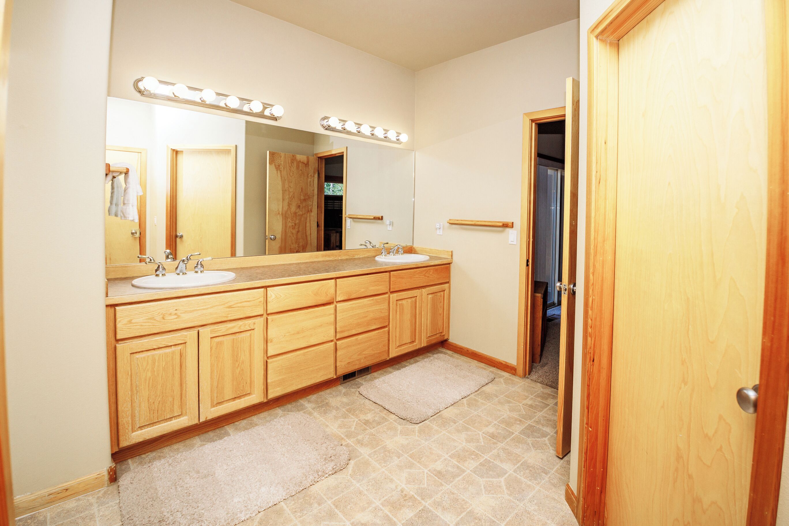 Une salle de bain avec un meuble-lavabo en bois, deux vasques, un grand miroir avec éclairage intégré et une porte donnant sur une autre pièce.