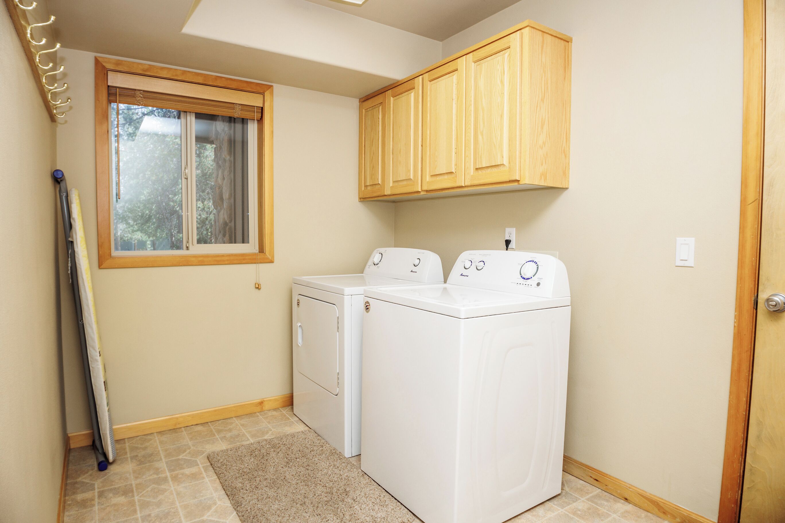 A laundry room with two white washers and dryers, wooden cabinets above, a window with blinds, and a door on the right.