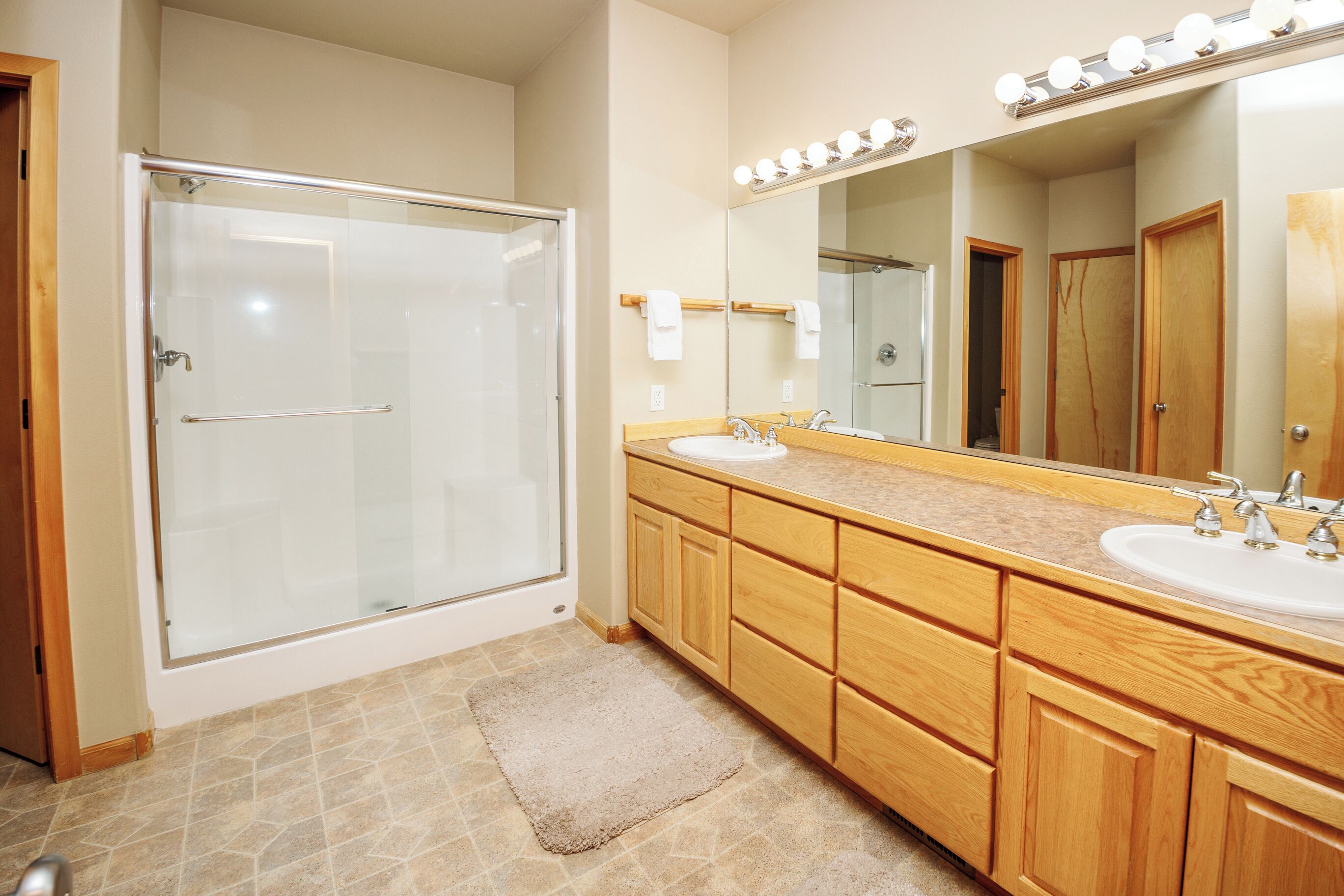 Ein Badezimmer mit einer Glaskabine zur Dusche, einem Holzmöbel zur Ablage mit zwei Waschbecken und einem großen Spiegel mit Beleuchtung darüber.