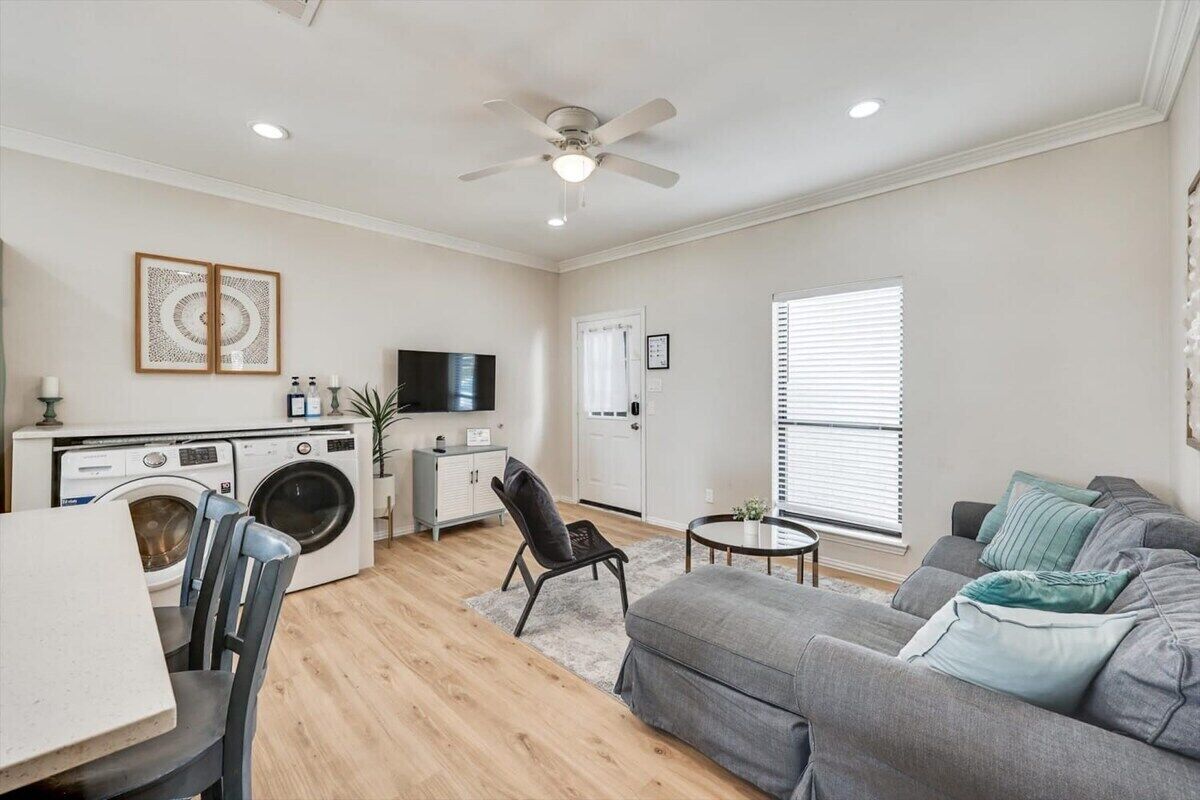 A living room with a grey sofa, a small round table, a washing machine, a ceiling fan, and a TV.