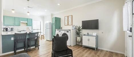 A modern kitchen with a dining area, a living room with a sofa, and a TV mounted on the wall.