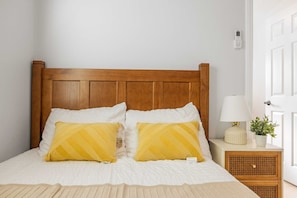 Una cama bien hecha con tablero de madera, dos almohadas con diseño amarillo, una lámpara de noche y una pequeña planta en maceta sobre una mesita.