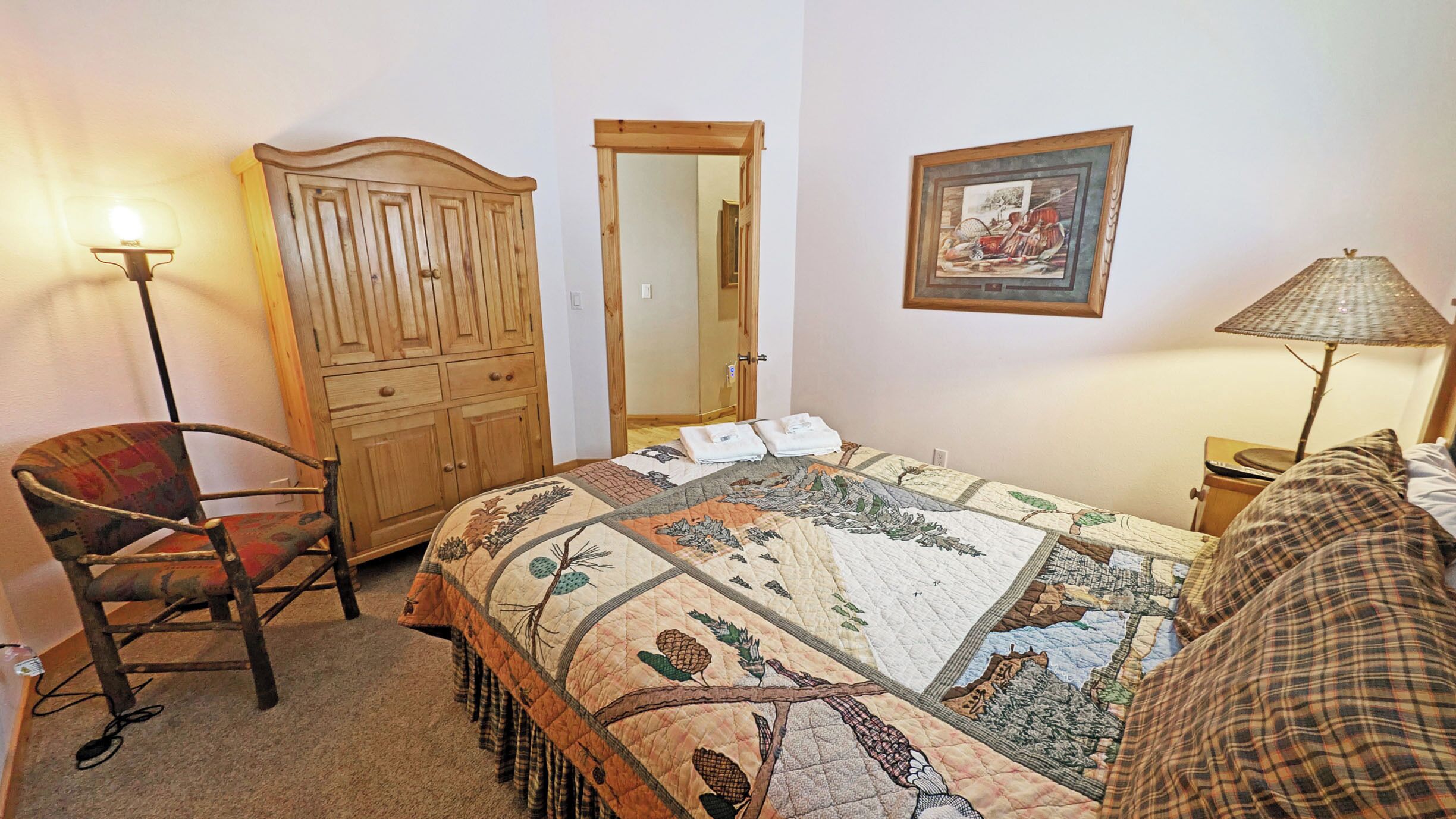 A bedroom with a bed, a wooden wardrobe, a chair, a bedside table with a lamp, and a framed picture on the wall.