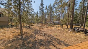 A forested backyard with a wooden deck, surrounded by tall pine trees and a house in the background. - Whistling Swan 8 (Bend)