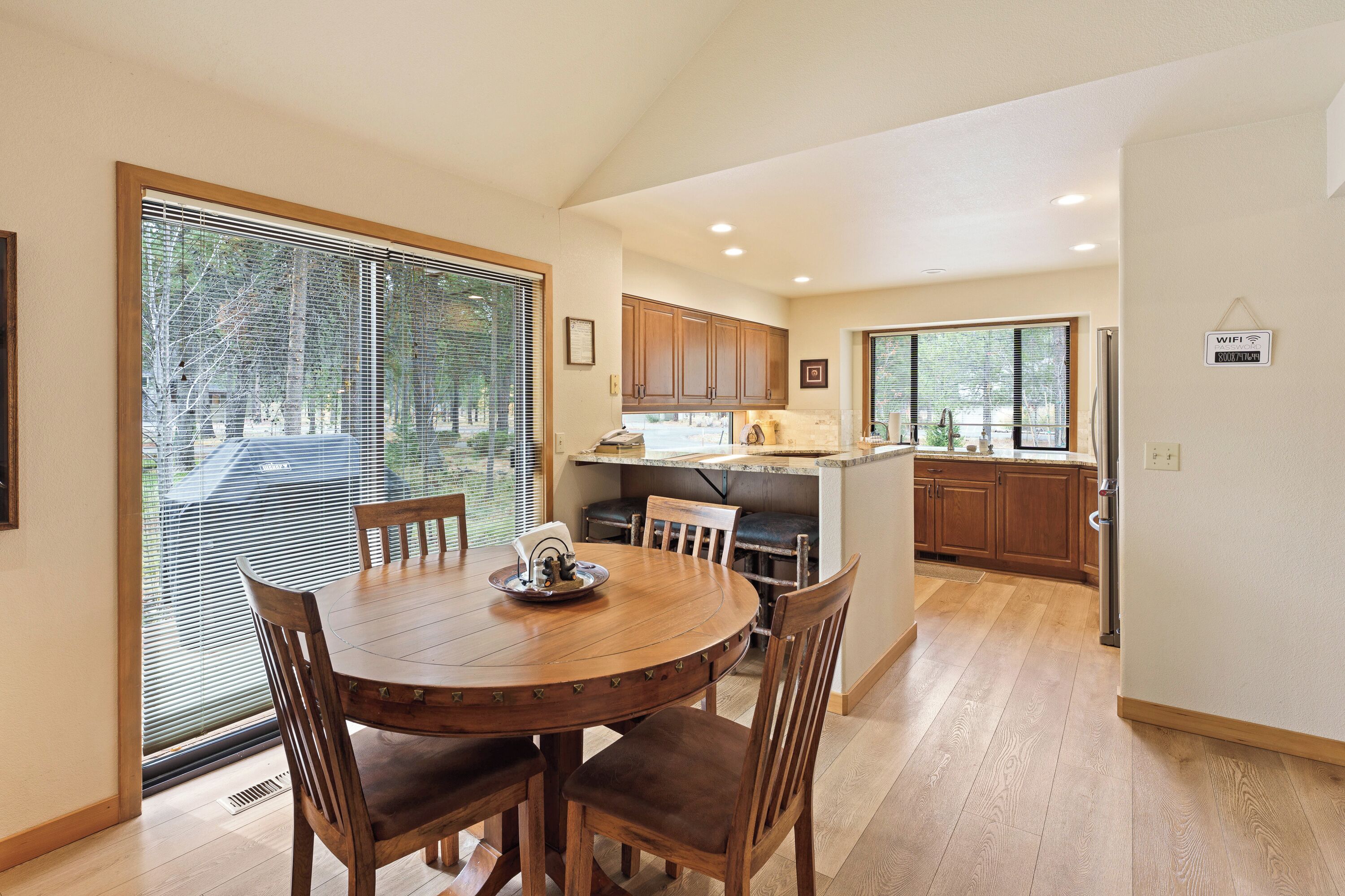 A dining area with a round wooden table and chairs, a kitchen with wooden cabinets, and a large window with blinds.