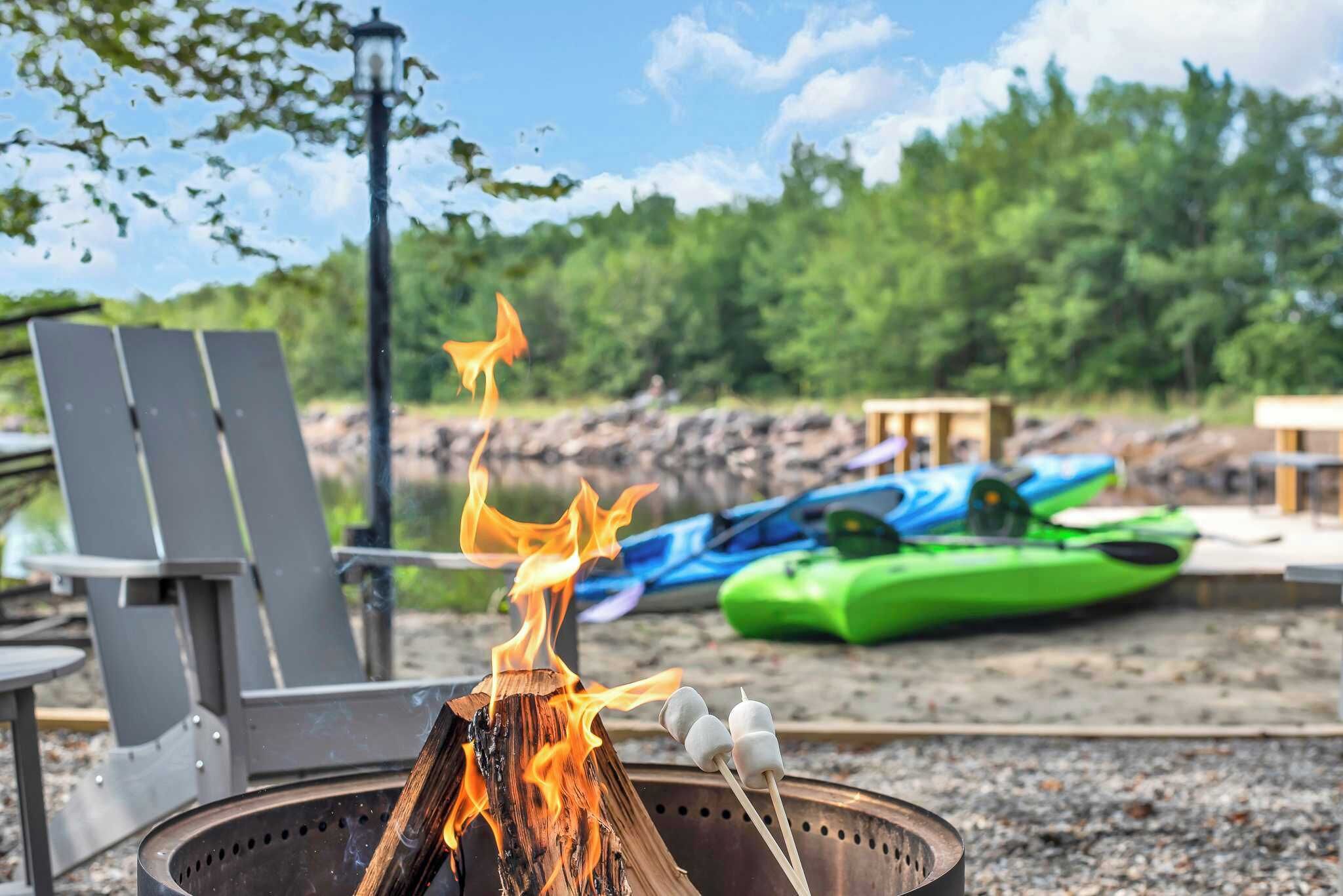 Ein Lagerfeuer mit Marshmallows, Gartenstühlen und Kajaks im Hintergrund.
