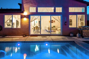 A modern house with a swimming pool, large glass doors leading to a living area, and outdoor lighting. - @ Marbella Lane - Desert Dream Pool/spa & Games (Desert Hot Springs)