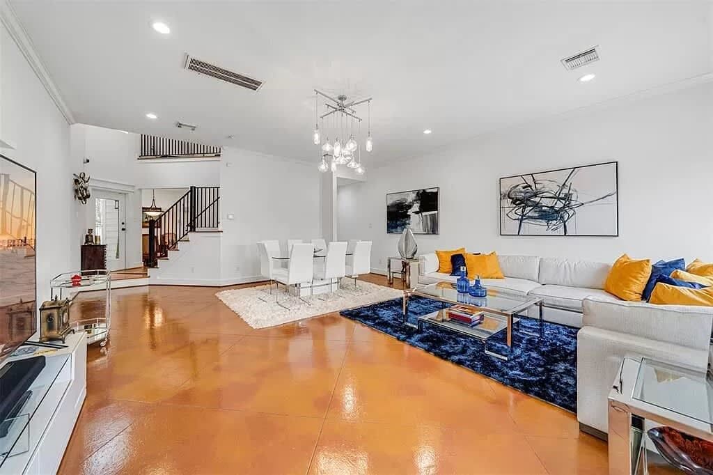 A spacious living room with a polished wooden floor, a white sofa with yellow cushions, a glass coffee table, and a modern chandelier.