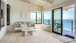 A spacious living room with a large window, a chandelier, a sofa with cushions, and a coffee table. - Oceanfront Neptune Ave Home | Panoramic Views, Luxury Coastal Living, Walk To The Beach (Encinitas)