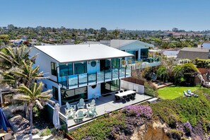 A modern two-story house with a glass-enclosed balcony, outdoor seating, and a landscaped garden. - Oceanfront Neptune Ave Home | Panoramic Views, Luxury Coastal Living, Walk To The Beach (Encinitas)
