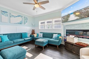 A living room with a teal sofa set, a ceiling fan, and a fireplace with a TV. - Jolly Rogers: Oceanfront Paradise with Pool, Sleeps 20, 7 bed/ 5.5 bath and two kitchens! (Oak Island)