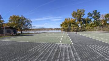 Un court de tennis en plein air, doté d’un filet, entouré d’arbres et avec une étendue d’eau en arrière-plan.