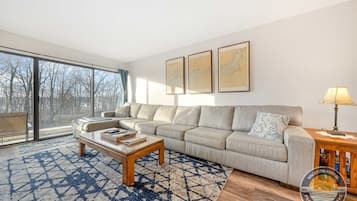 A modern living room with a sectional sofa, a coffee table, and a large window offering a view of trees.