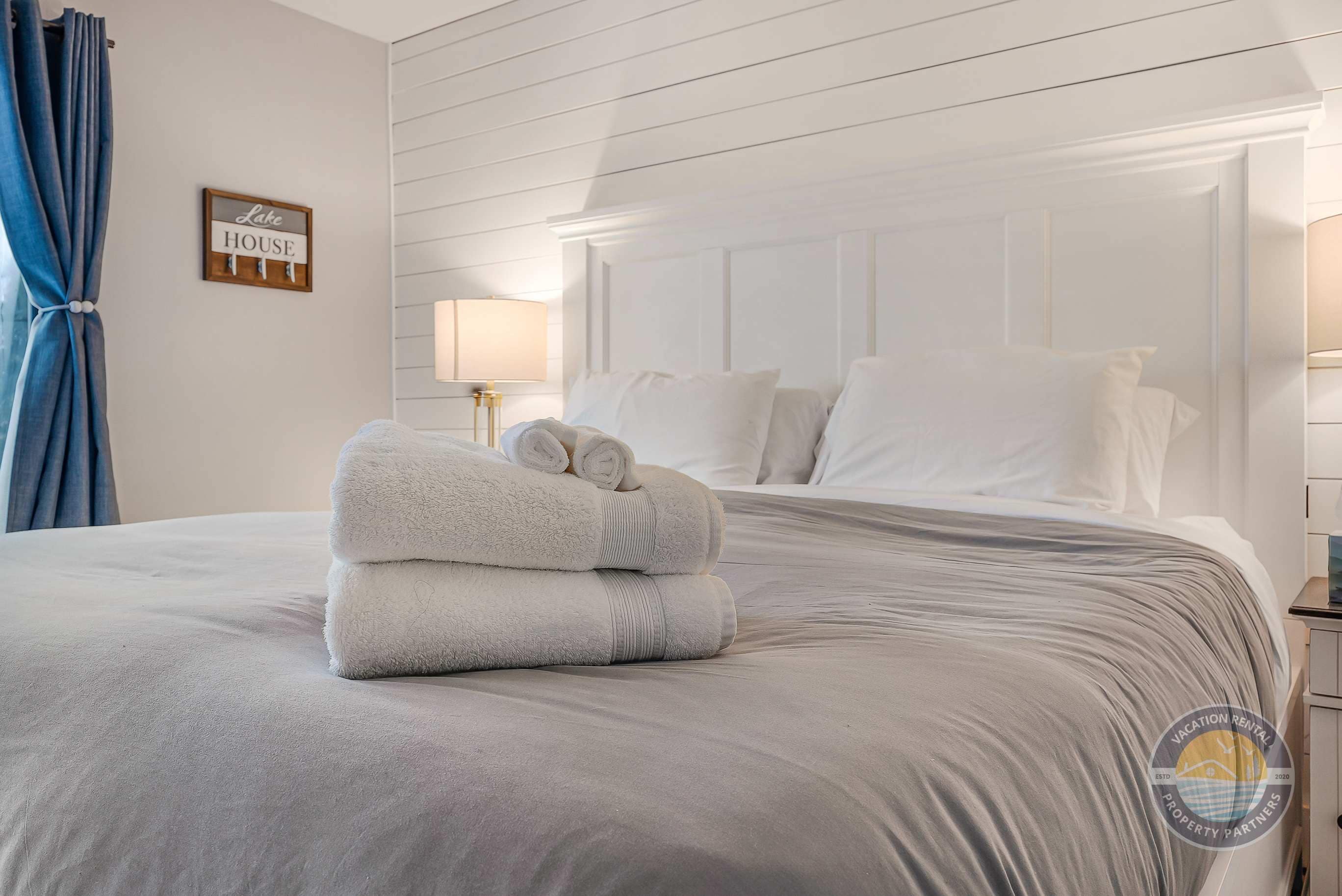 A neatly made bed with folded towels, a wall-mounted sign reading 'Lake House,' and a bedside lamp.