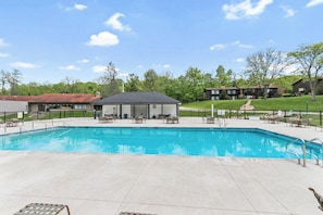A swimming pool with lounge chairs and a small building, surrounded by a fence and trees.