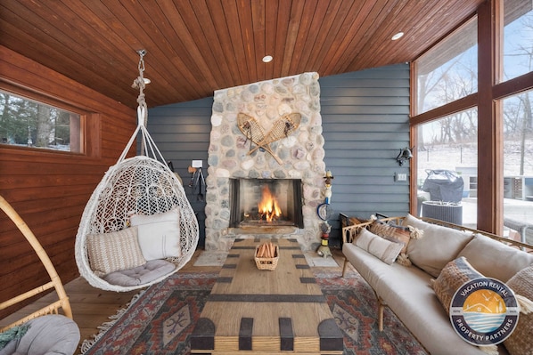 A cozy interior with a stone fireplace, a hanging chair, a sofa, and a view of the outdoors.