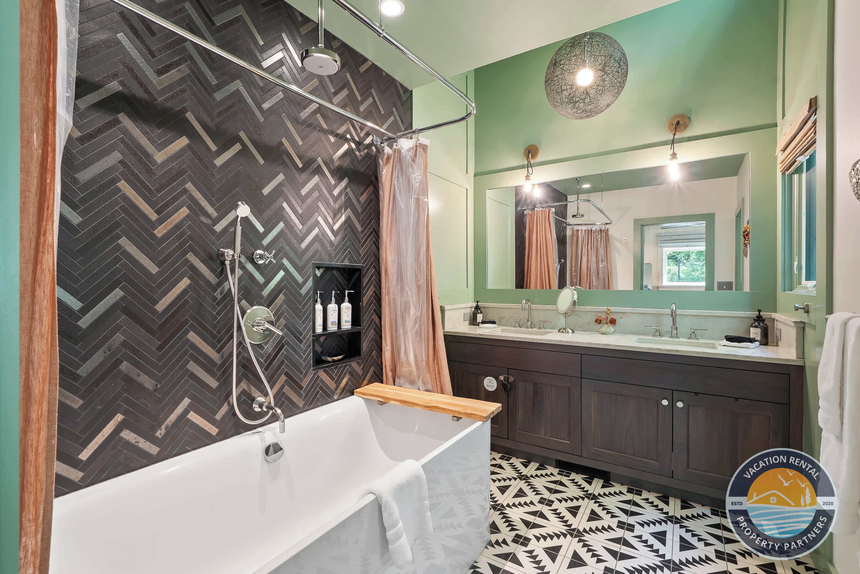 Un bagno moderno con parete a motivo a spina di pesce, vasca freestanding, mobile lavabo con specchio e pavimento a motivi geometrici.