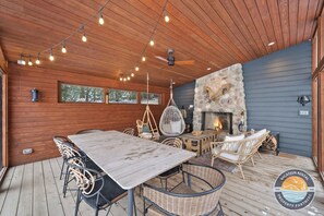A wooden deck with a dining table and chairs, a hanging chair, and a stone fireplace.