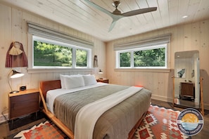 A bedroom with a bed, nightstands, a ceiling fan, large windows, and a rug.