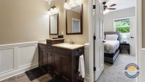 Un baño con tocador, lavamanos y dos espejos. Se ve una puerta que da a un dormitorio con cama y mesita de noche.